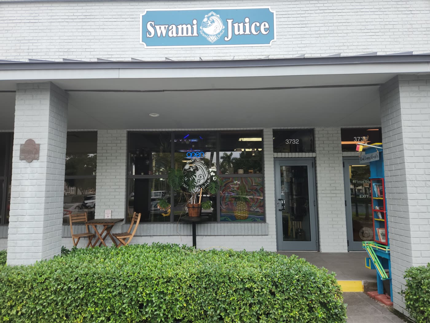 The Swami Juice storefront at 3732 Davie Rd — blue sign over a white brick building, with a bistro table under the walkway