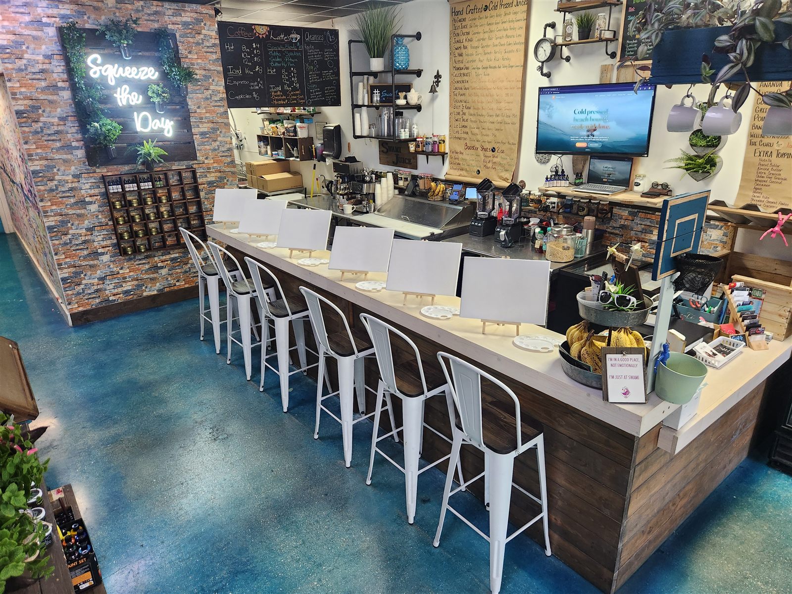 The bar at Swami Juice set for a paint night — blank canvases on easels and palettes lined up in front of the white stools, under the Squeeze the Day neon